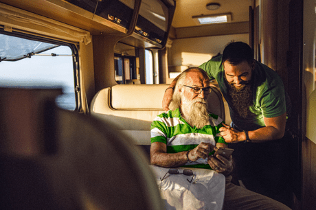 Two people in a campervan, one sitting looking at a smartphone, the other standing nearby offering help.