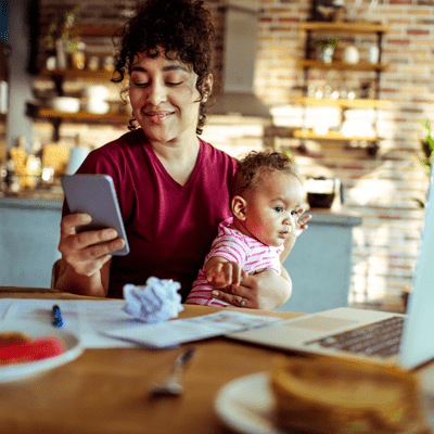 A woman sitting at a table holding a smart phone with a baby on her lap