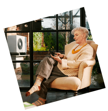 Person sitting in a cosy armchair near large windows, holding a thermostat. A heat pump is visible outside the window.