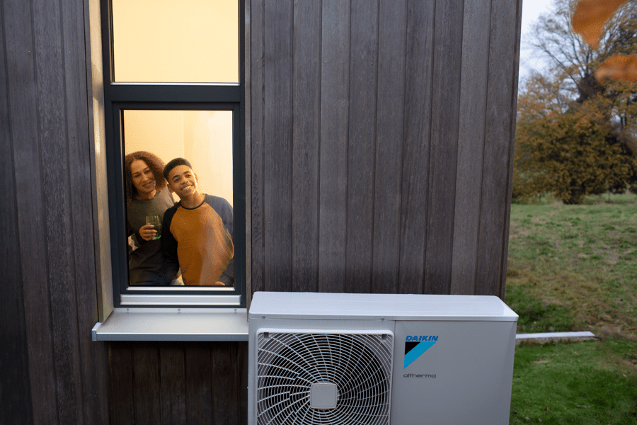 A woman and a teenager look smiling out of a window. There is a heat pump outside. 