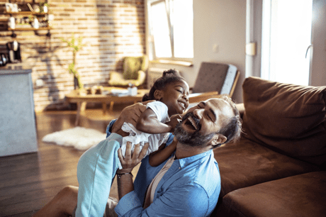 A man lifting his young daughter and laughing in his living room on a bright, sunny day