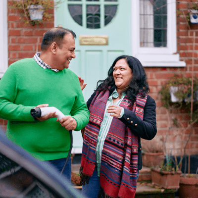 A man holding an EV charger outside a house, chatting to a woman, with a car in the foreground