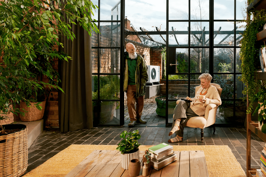 An older couple in their home with heat pump in background. The woman sits on the chair while the man stands.