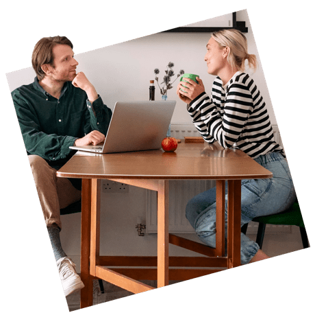 Couple sat at table having a conversation whilst browsing on laptop